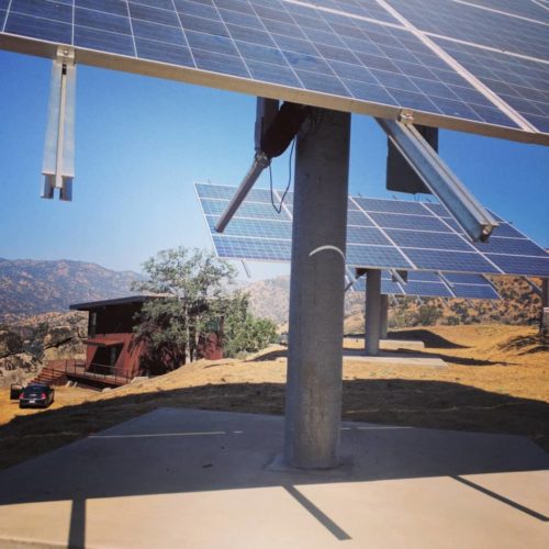 Large solar panels mounted on metal frames over concrete ground, with a small red building and mountains in the background under a clear sky.