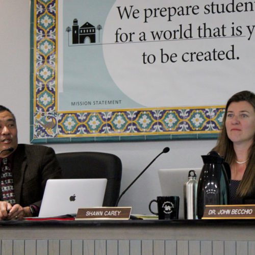 Two school board members, one asian man and one caucasian woman, sitting at a desk during a meeting, with a mission statement on the wall behind them.