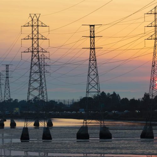 Sunset view over a river with electrical power lines and pylons extending across the landscape, an airplane visible in the sky.