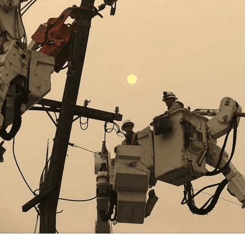 Utility workers in bucket lifts repair power lines against a hazy, smoke-filled sky with a dimly visible sun.