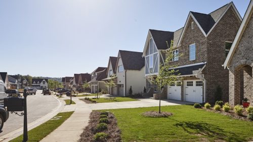 A sunny suburban street lined with modern two-story brick houses featuring front garages and neatly manicured lawns.