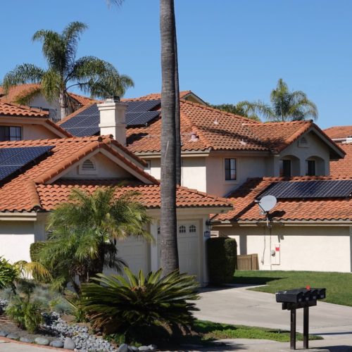 Suburban street view depicting two houses with solar panels on red tile roofs, clear blue sky, and a tall palm tree.