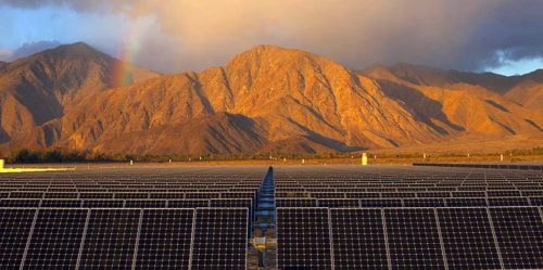 Solar panels in the foreground with a rainbow and sunlit mountains in the background under a cloudy sky.