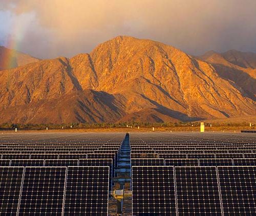 Solar panels in the foreground with a rainbow and sunlit mountains in the background under a cloudy sky.