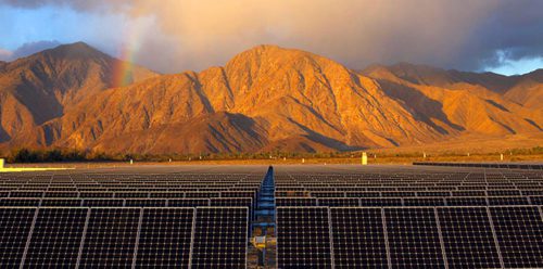 Solar panels in the foreground with a rainbow and sunlit mountains in the background at sunset.