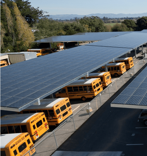 Aerial view of school buses parked under large solar panel installations at a bus depot.