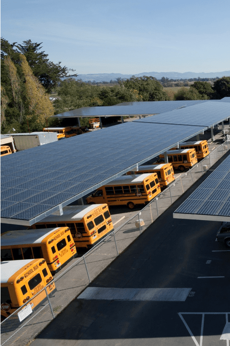 Aerial view of school buses parked under large solar panel installations at a bus depot.