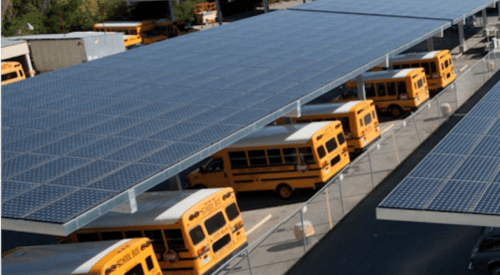 Yellow school buses parked under a large solar panel canopy in a sunny parking lot.