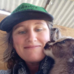 A woman in a hat and jacket kissing a baby animal.